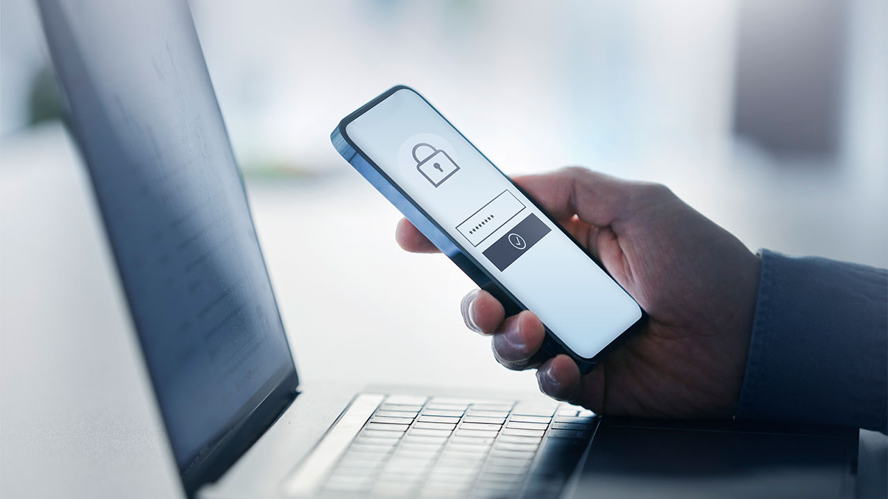Person hält Smartphone mit geschütztem Login-Fenster über einen Laptop.